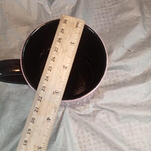 Puede incluir: Una taza de caf&eacute; negra con borde y asa rosa. Una regla de madera se coloca dentro de la taza, mostrando un di&aacute;metro de aproximadamente 7,6 cm. El interior de la taza es negro.