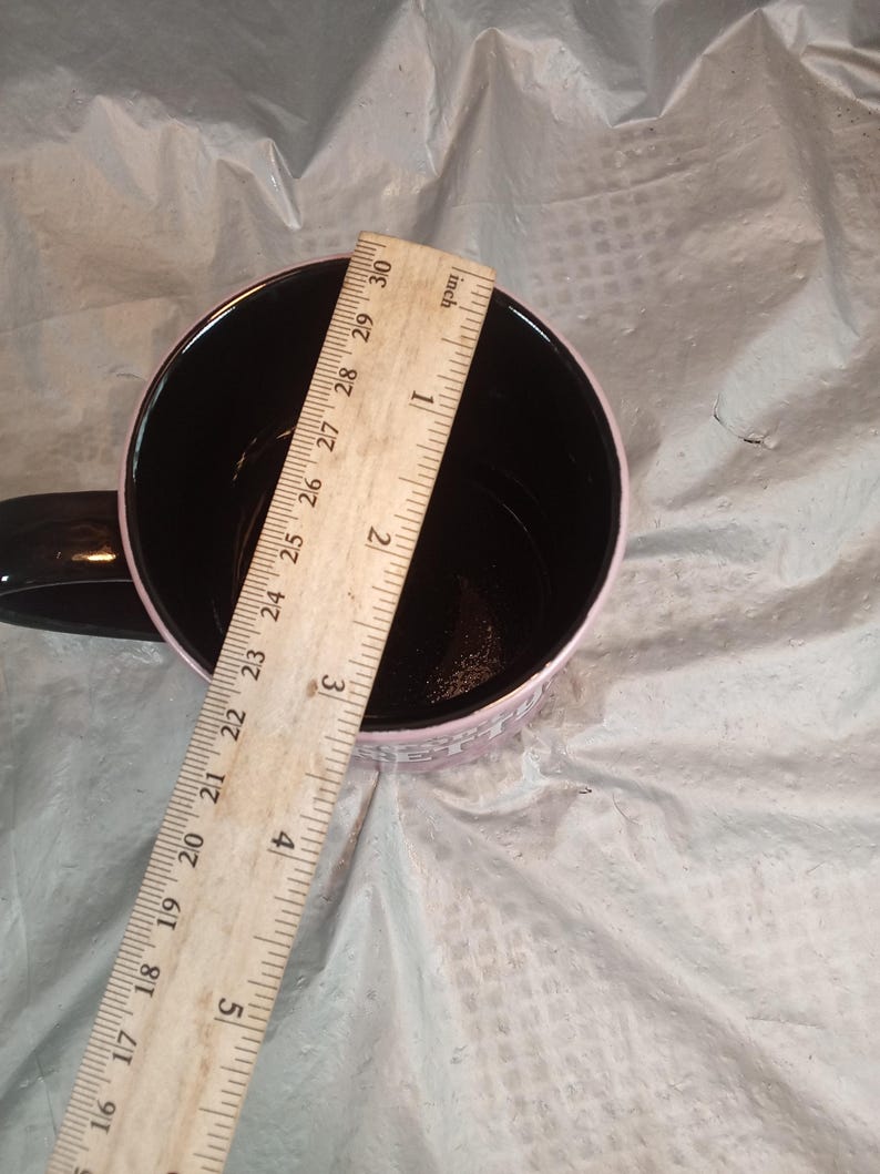 Puede incluir: Una taza de caf&eacute; negra con borde y asa rosa. Una regla de madera se coloca dentro de la taza, mostrando un di&aacute;metro de aproximadamente 7,6 cm. El interior de la taza es negro.
