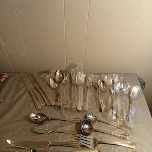 May include: A selection of silver-coloured cutlery, including forks, spoons, knives, and serving utensils. Some pieces are individually wrapped in plastic. The cutlery is arranged on a grey surface, suggesting a dining or serving set.