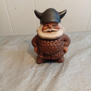 May include: A ceramic Viking figurine with a black horned helmet, a white beard, and a brown tunic. The figurine is smiling and has its hands on its hips. The texture of the tunic is visible.