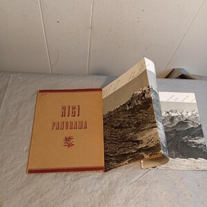 May include: A vintage book titled "Rigi Panorama" with a brown cover and red trim. The book is open, revealing black and white landscape photographs of mountains. The cover features the title in red and a small floral design.