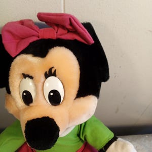 May include: A plush toy featuring a cartoon character with a black head, large white eyes, and a black nose. The character wears a pink bow, a green shirt, and a red skirt. The toy is made of soft, plush material.