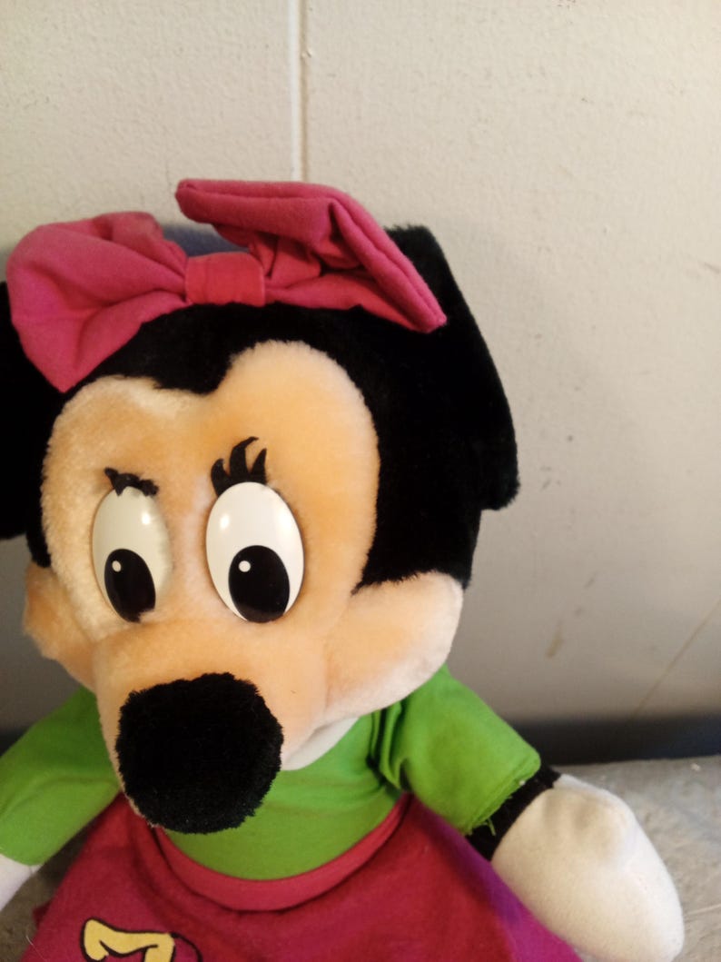 May include: A plush toy featuring a cartoon character with a black head, large white eyes, and a black nose. The character wears a pink bow, a green shirt, and a red skirt. The toy is made of soft, plush material.