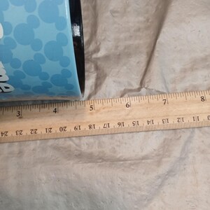 Pu&ograve; includere: Un bicchiere blu con bordo nero e sfondo azzurro con forme di testa di Topolino bianche. Il bicchiere &egrave; accanto a un righello di legno. Il bicchiere ha un diametro di circa 10 cm.