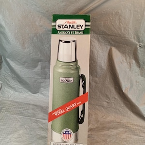 May include: A green Stanley Aladdin steel quart bottle in its original packaging. The box features the text Aladdin Stanley America's #1 Brand, Steel Quart Bottle, and Guaranteed for Life. The bottle has a silver cap and a black side handle.