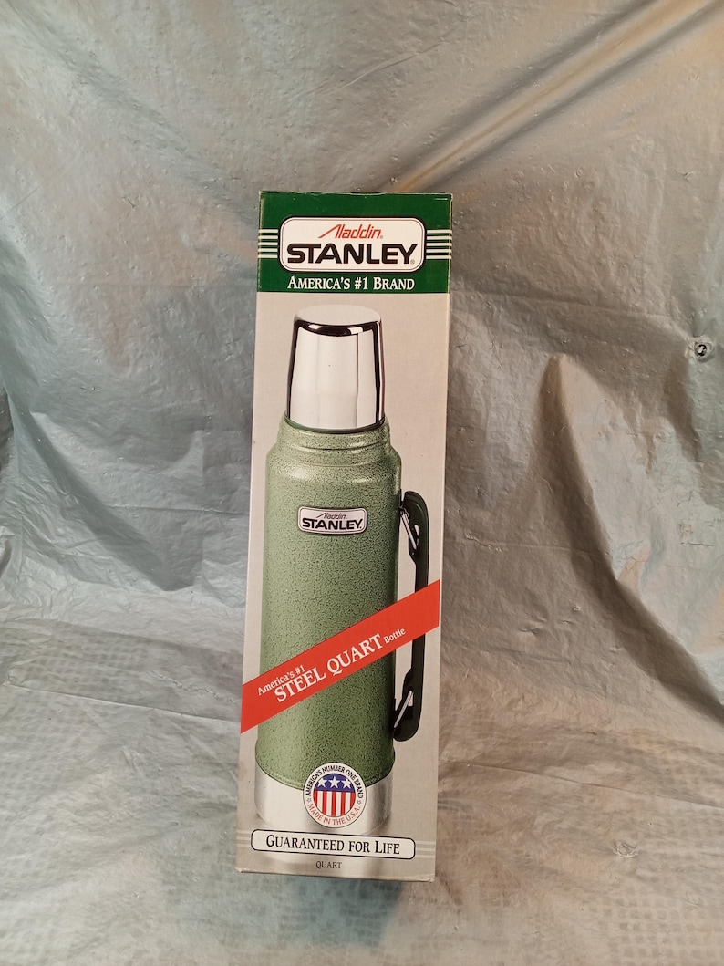 May include: A green Stanley Aladdin steel quart bottle in its original packaging. The box features the text Aladdin Stanley America's #1 Brand, Steel Quart Bottle, and Guaranteed for Life. The bottle has a silver cap and a black side handle.