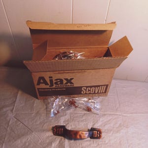 May include: A cardboard box labeled "Ajax Hardware Housing Products Scovill" with an open lid. Inside are several hardware items in plastic packaging. A bronze-colored cabinet pull is in the foreground.