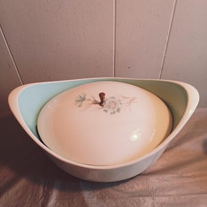 以下が含まれることがあります： 薄い青色の縁取りと、それに合わせた蓋が付いた白いセラミック製の食器。蓋には花柄と木製のつまみがある。