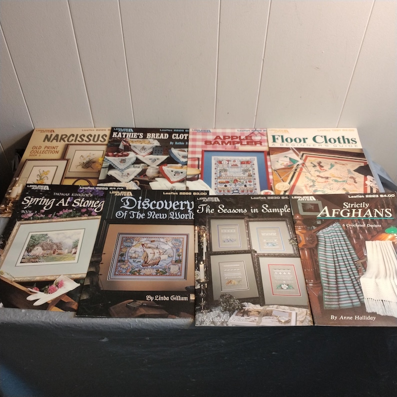Pode incluir: Oito livros de padr&otilde;es de ponto cruz com capas coloridas. Os t&iacute;tulos incluem "Narcissus Old Print Collection", "Kathie's Bread Cloths", "Apple Bampler", "Floor Cloths", "Spring At Stoneg", "Discovery of the New World", "The Seasons in Sample" e "Strictly Afghans".