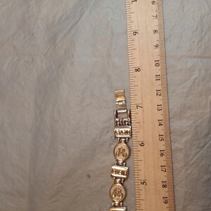 May include: A silver-toned metal link bracelet featuring alternating rectangular links with the word MICKEY and circular links with a Mickey Mouse head icon, positioned next to a wooden ruler showing a length of approximately 19 cm.