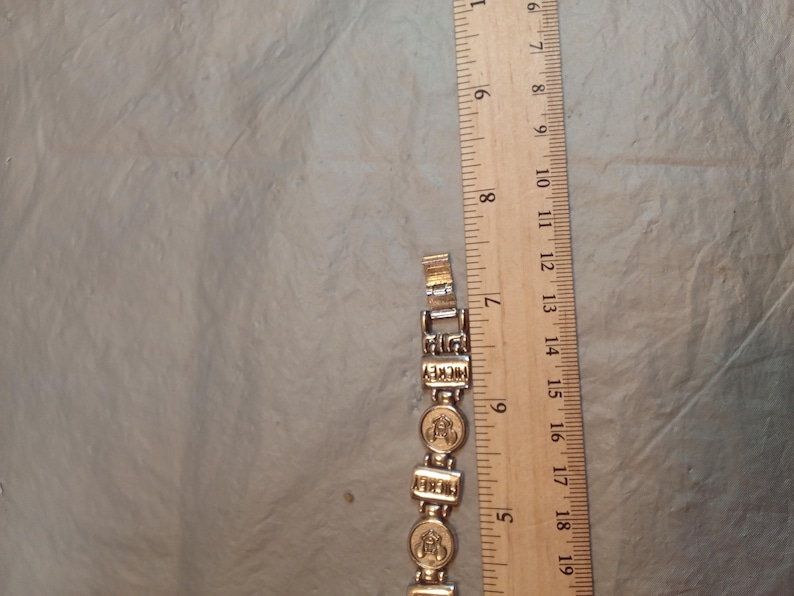 May include: A silver-toned metal link bracelet featuring alternating rectangular links with the word MICKEY and circular links with a Mickey Mouse head icon, positioned next to a wooden ruler showing a length of approximately 19 cm.