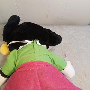 May include: A plush doll with black ears, a green top, and a pink skirt. The doll has white gloves and a yellow pom-pom. The doll is lying on a light gray surface.