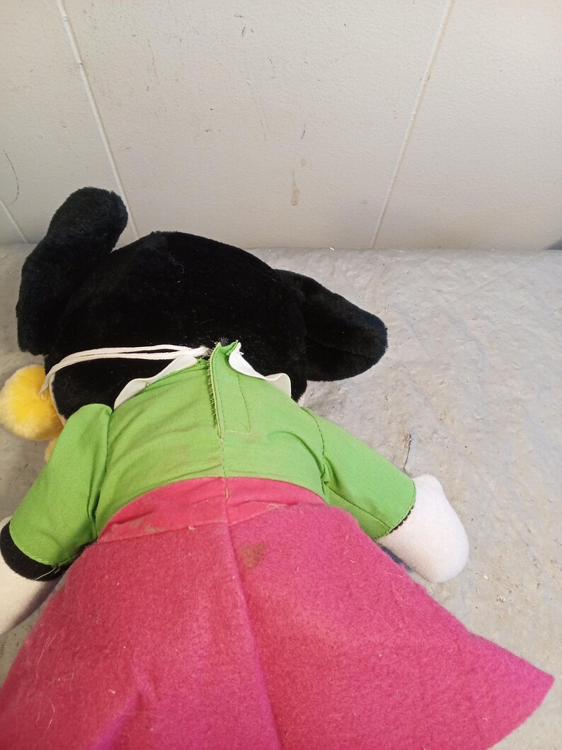 May include: A plush doll with black ears, a green top, and a pink skirt. The doll has white gloves and a yellow pom-pom. The doll is lying on a light gray surface.