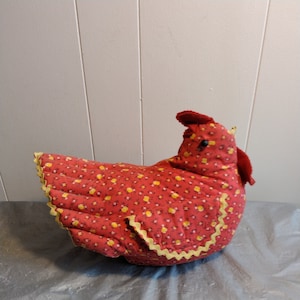 May include: A stuffed chicken-shaped decorative item. The chicken is red with a yellow floral pattern and yellow trim. It has a red comb and wattle, and black eyes. The item is likely a kitchen or home decor piece.