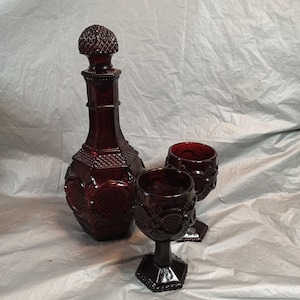 May include: A ruby red glass decanter with a matching stopper and two stemmed goblets. The decanter and goblets have an ornate, textured design. The decanter is approximately 30 cm tall. The goblets are approximately 13 cm tall.