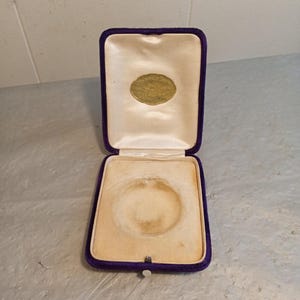 Puede incluir: Caja de joyería vintage abierta, con exterior de terciopelo morado e interior de satén color crema. La caja revela una hendidura circular y un medallón dorado. La caja está diseñada para guardar una joya.