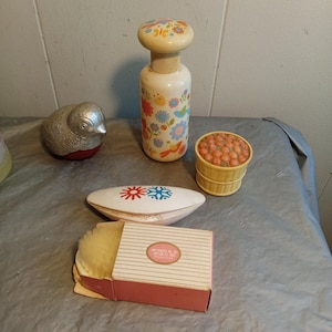 May include: A collection of vintage cosmetic items, including a floral-decorated bottle, a silver bird-shaped container, a small basket filled with orange spheres, a white and red container, and a powder box with a pink label.