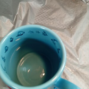 May include: A light blue ceramic mug with a handle. The interior and handle are a lighter shade of blue. The rim of the mug features small, dark blue illustrations of sea creatures. A red fish is visible on the exterior.