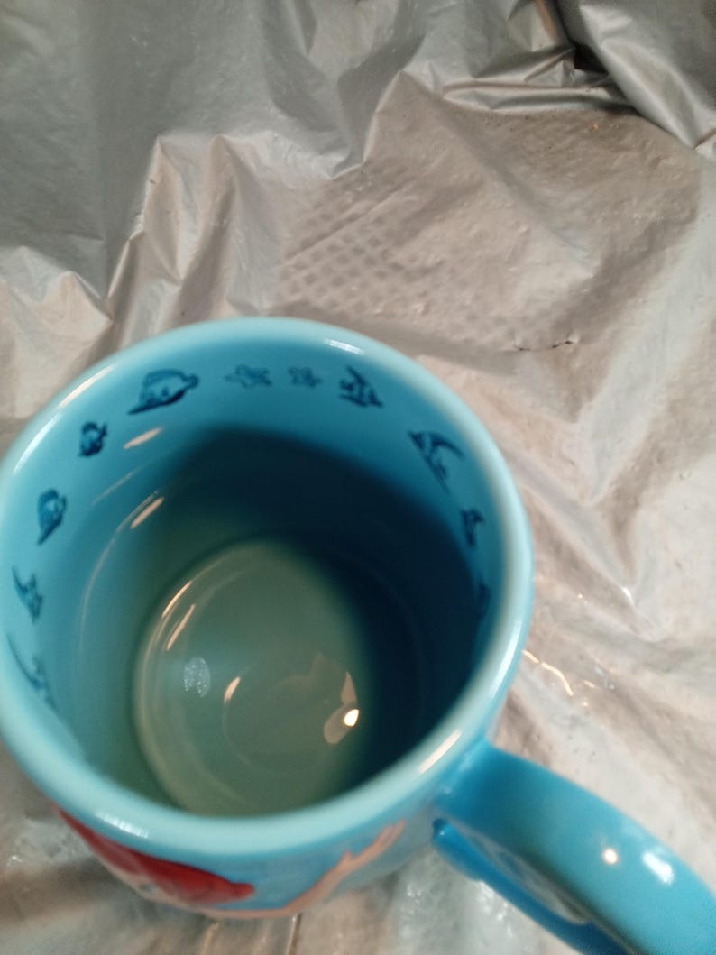 May include: A light blue ceramic mug with a handle. The interior and handle are a lighter shade of blue. The rim of the mug features small, dark blue illustrations of sea creatures. A red fish is visible on the exterior.