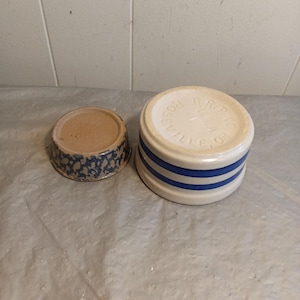May include: Two ceramic containers. One is a small, round bowl with a brown interior and a blue and white speckled exterior. The other is a larger, cylindrical container with a white base and two blue stripes. The top of the larger container has the text "ROSEVILLE, OHIO".