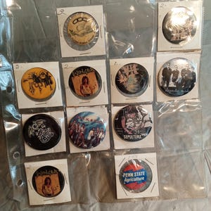 May include: A collection of vintage pinback buttons featuring band logos and other designs. The buttons are in a clear plastic sheet. Some buttons have text that reads "Winger", "Sepultura", and "Penn State Agriculture". Other buttons feature images of bands.