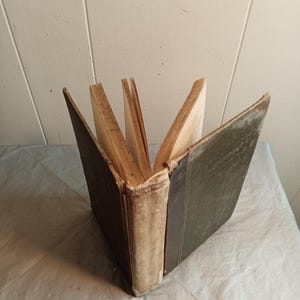 May include: An open antique book with aged, yellowed pages. The spine is off-white with a dark green cover on the right side and a dark brown cover on the left. The book is fanned open, displaying the pages.