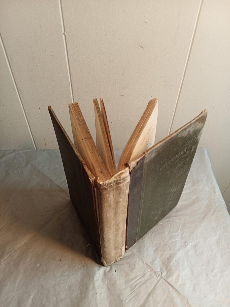 May include: An open antique book with aged, yellowed pages. The spine is off-white with a dark green cover on the right side and a dark brown cover on the left. The book is fanned open, displaying the pages.