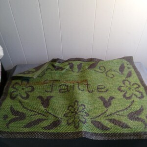 May include: Green and brown doormat with the word "faite" woven into the design. The doormat has a floral pattern with leaves and flowers.