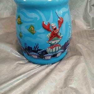 May include: A light blue ceramic mug featuring an underwater scene with cartoon fish and coral. The mug has a design of Sebastian the crab from The Little Mermaid. The mug is cylindrical with a slightly flared top.