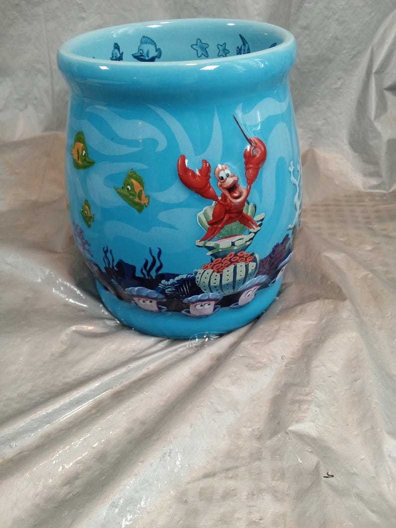 May include: A light blue ceramic mug featuring an underwater scene with cartoon fish and coral. The mug has a design of Sebastian the crab from The Little Mermaid. The mug is cylindrical with a slightly flared top.
