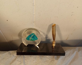Könnte beinhalten: Ein Schreibtischset mit schwarzem Marmorsockel. Es enthält eine transparente Acrylkugel mit einem türkisblauen Mineral und einen goldfarbenen Stifthalter. Das Mineral hat ein kleines Etikett. Das Set wird auf einer hellgrauen Oberfläche präsentiert.