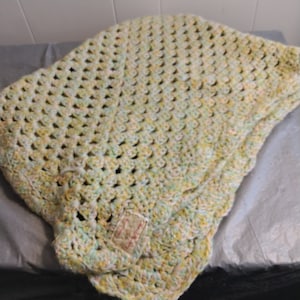 以下が含まれることがあります： 黄色、緑、白の糸で編まれた、赤ちゃん用の毛布です。毛布はフリルのある縁取りになっており、小さなタグには「The Village Knitter」と書いてあります。