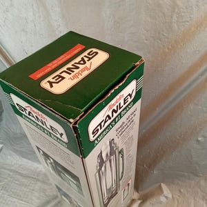 May include: A green and white cardboard box for a Stanley Aladdin vacuum bottle. The packaging features the text Stanley Aladdin, America's #1 Brand, and states the capacity is 1 quart/950 ml. The box displays a graphic of the steel bottle.