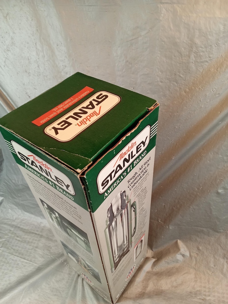 May include: A green and white cardboard box for a Stanley Aladdin vacuum bottle. The packaging features the text Stanley Aladdin, America's #1 Brand, and states the capacity is 1 quart/950 ml. The box displays a graphic of the steel bottle.