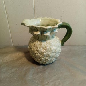 May include: A ceramic pitcher shaped like a pineapple. The pitcher is primarily a light green color with a textured surface, a green handle, and a ruffled rim. The pitcher is a decorative item.
