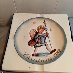 Può includere: Un piatto natalizio decorativo del 1982 con un angelo dipinto con uno strumento musicale e un bastone. Il piatto ha un bordo blu con stelle dorate e le parole "Christmas 1982". Il piatto è nella sua scatola originale.