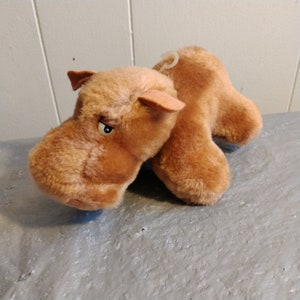 Può includere: Un giocattolo di ippopotamo di peluche marrone con un'espressione scontrosa. Il giocattolo ha un anello sul retro.