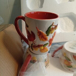 May include: A ceramic mug with a red interior and handle, decorated with autumn leaves in shades of orange, yellow, and green. The mug is white with a glossy finish and is part of a set. The mug is in its original packaging.