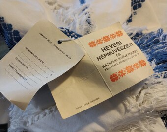 May include: A white tag with black text that reads "HEVESI NÉPMŰVÉSZETI ES HAXIIPARI SZÖVETKEZET 3360 HEVES, KOSSUTH L. UT 26-28" and a smaller black text that reads "OKISZ LABOR NYOMDA". The tag is attached to a white fabric with blue embroidery.