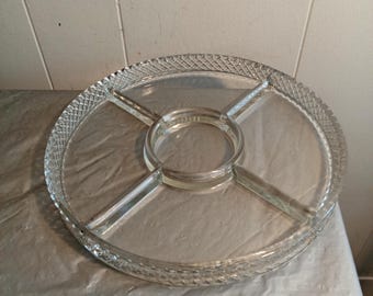 May include: A clear glass serving tray with a circular shape and four divided sections. The rim features a textured, diamond-patterned design. The center has a small, raised circular area. Ideal for serving snacks or appetizers.