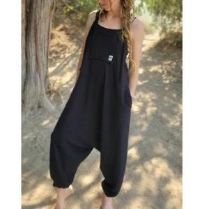 May include: A solid black sleeveless jumpsuit featuring a loose, relaxed fit with a low-hanging crotch area, thin shoulder straps, and a square-shaped patch pocket on the chest. The garment has a simple, unadorned design with cuffed leg openings.