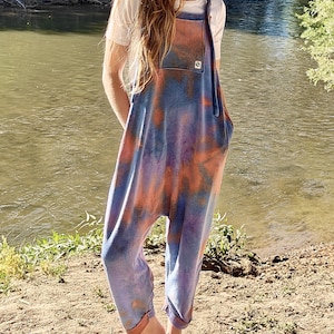 May include: A tie-dye jumpsuit in shades of blue, orange, and purple. The jumpsuit has a relaxed fit with a bib-style top, large front pockets, and rolled cuffs. The model is wearing a white t-shirt underneath.