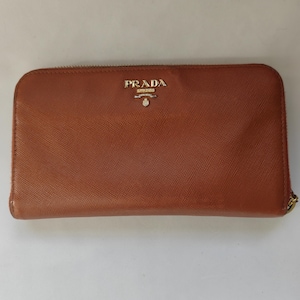 May include: A rectangular brown wallet with a textured surface and a gold-tone zipper closure. The front features the gold-colored Prada Milano logo with a crest emblem centered near the top edge. The wallet has rounded corners and a structured design.