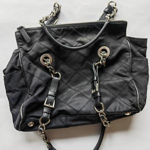 May include: Black quilted tote bag with silver-tone chain straps and accents. The bag features a diamond pattern, a top zipper closure, and leather details. It has two handles and a shoulder strap.