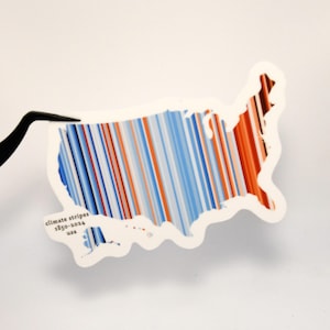 May include: A sticker in the shape of the United States with a climate stripes design. The stripes are vertical and alternate between blue, white, and red. The text "climate stripes 1850-2024 usa" is printed on the sticker.