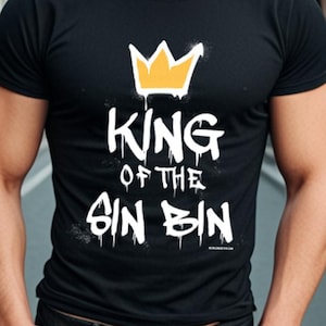 May include: Black crew neck t-shirt with the text "KING OF THE SIN BIN" in white and a yellow crown graphic. The t-shirt is paired with dark wash jeans. The person is smiling and standing outdoors.
