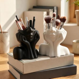 May include: Two ceramic desk organizers shaped like human heads resting on hands. One organizer is matte black and holds various pens and pencils, while the other is matte white and holds a collection of makeup brushes with brown bristles.