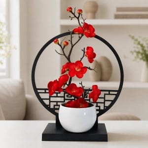 May include: A decorative arrangement featuring a white ceramic bowl containing artificial red plum blossoms on dark brown branches. The plant is framed by a black circular structure with a geometric lattice base, all mounted on a black rectangular stand.