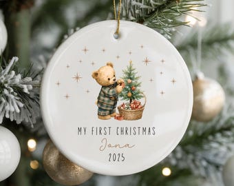 May include: A white ceramic Christmas ornament with a gold string, featuring a teddy bear decorating a Christmas tree. The text "MY FIRST CHRISTMAS Jana 2025" is printed on the ornament. The ornament is hanging on a Christmas tree with white and gold ornaments.