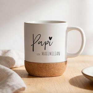 Könnte beinhalten: Weißer Keramikbecher mit Korkboden. Der Becher trägt das Wort "Papi" in Schreibschrift, ein kleines Herz und den Text "von Maximilian" in einer serifenlosen Schriftart. Der Becher hat einen weißen Henkel und steht auf einer Holzoberfläche.